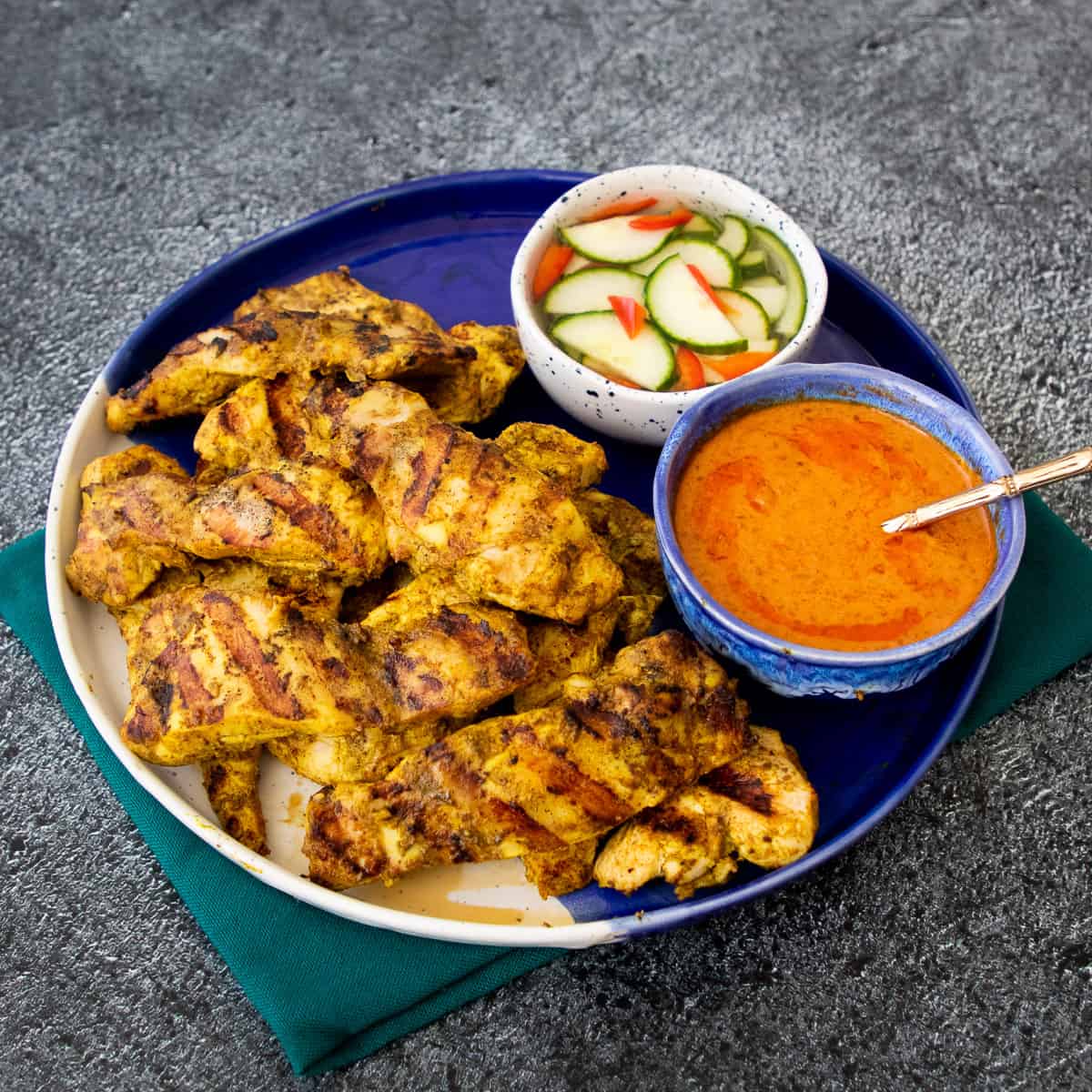 A plate of chicken satay with a bowl peanut sauce and a bowl cucumber pickle.